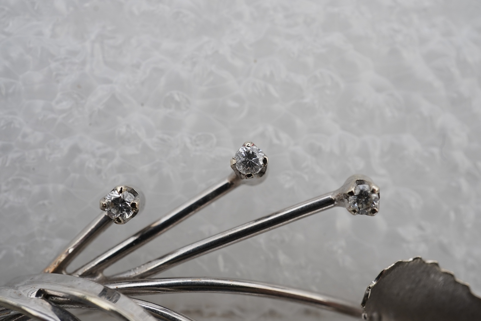 A diamond brooch, designed as stylised foliate spray in white gold, accented with three brilliant-cut diamonds totalling approximately 0.15 carats, length 5.2cm, stamped 750, gross weight 5 grams.